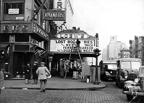 A man on a ladder changes the marquee lettering of the Loew’s theater on the corner of Second Avenue and 6th Street on the Lower East Side of Manhattan.  Several cars and trucks line the streets, two men in suits and hats stand together on the corner and a man with his back to us crosses 6th street.  The scene is very busy. 